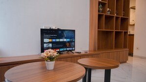 Una sala de estar moderna con un televisor de pantalla plana sobre un soporte de madera, dos mesas redondas de madera, un pequeño jarrón blanco con flores y un mueble de estanterías de madera contra la pared.