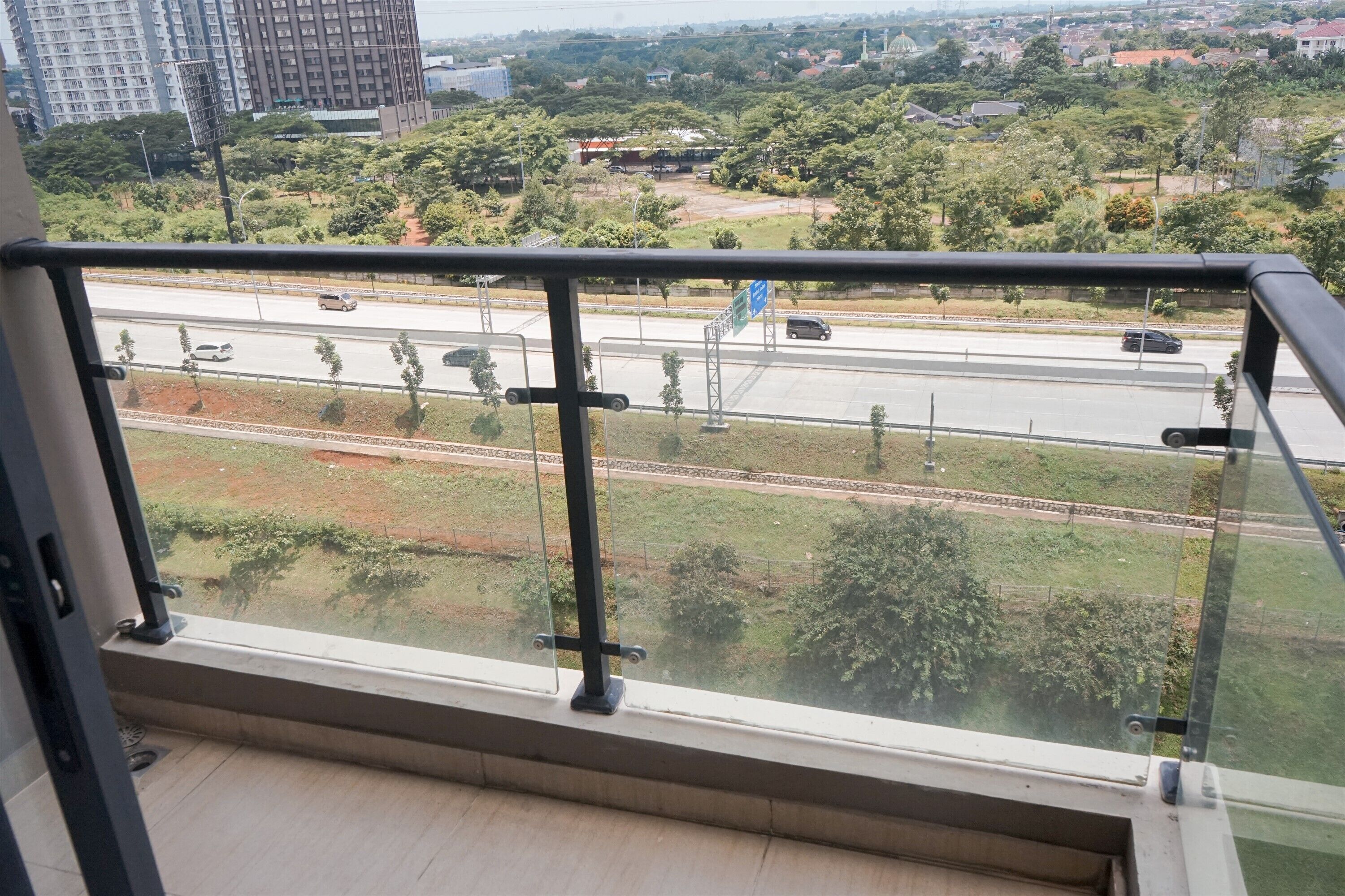 A view from a balcony with a metal railing, overlooking a road with vehicles, greenery, and distant buildings.