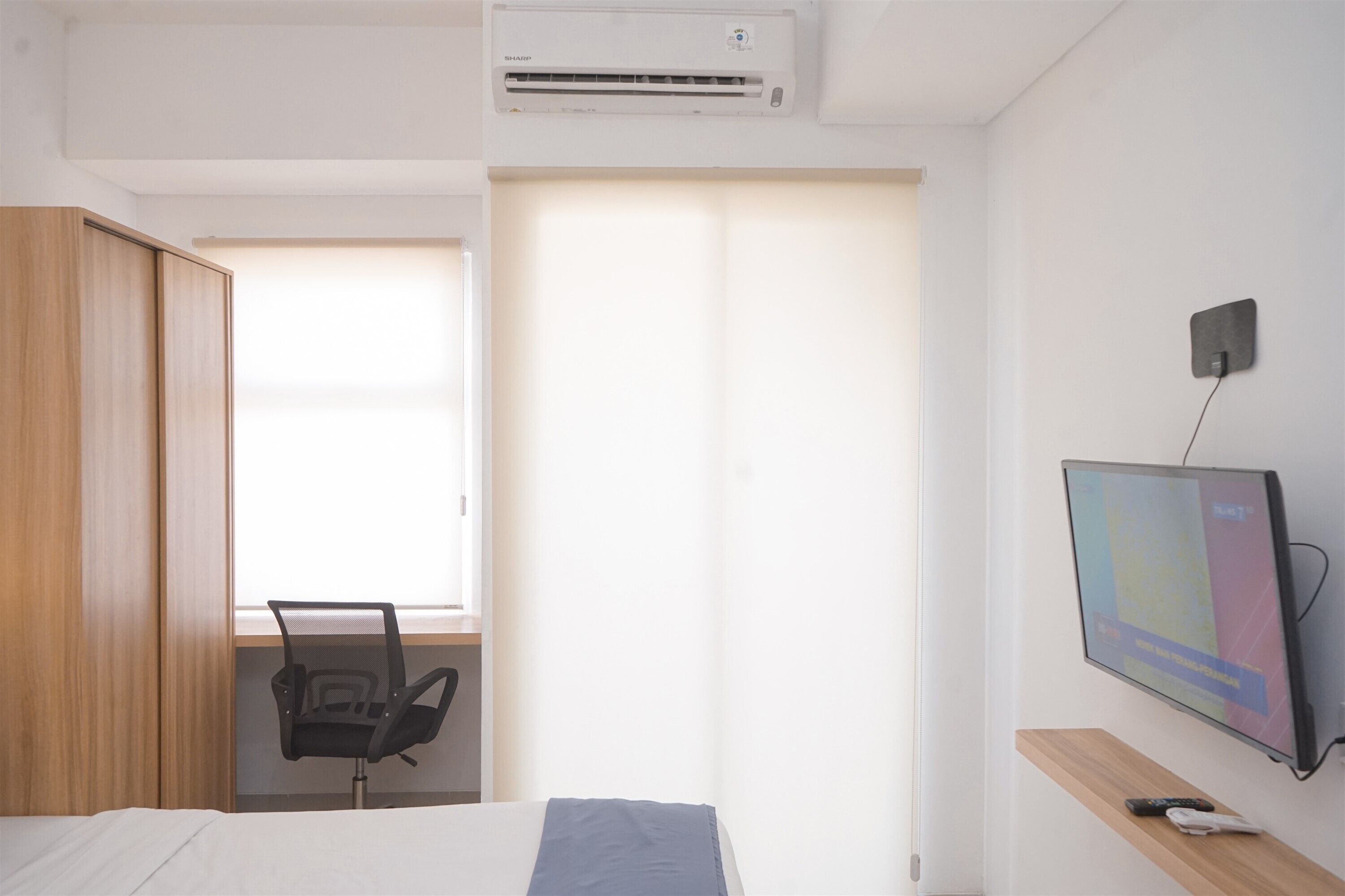 A hotel room with a bed, a desk with a chair, a TV mounted on the wall, and an air conditioning unit.