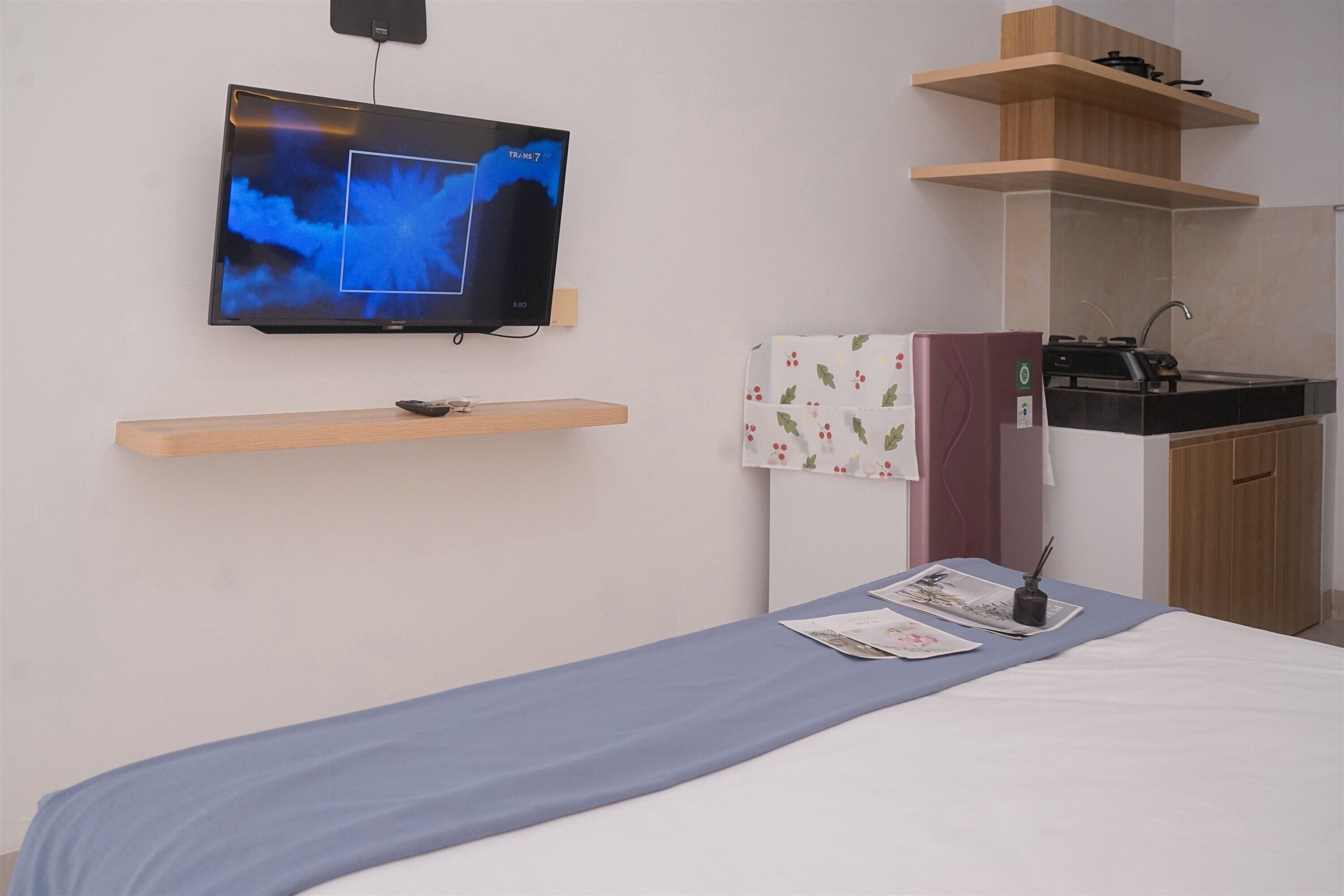 A hotel room with a bed, a TV mounted on the wall, a small kitchen area with a sink and stove, and a wooden shelf above the bed.