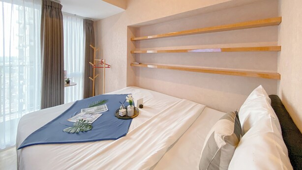 Une chambre d’hôtel avec un lit, une fenêtre avec des rideaux, une étagère en bois et une petite table sur laquelle se trouvent un vase et une plante.