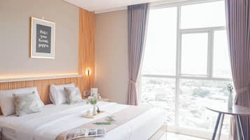 A hotel room with a large bed, a bedside table, a nightstand, a framed inspirational quote on the wall, and a view of the city through the window.
