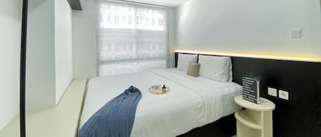 A modern hotel room with a bed, bedside table, and a window with blinds.