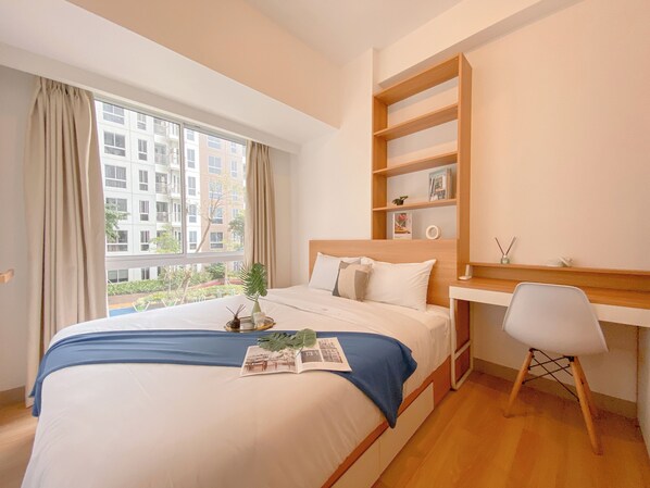 A modern bedroom with a large bed, a desk, and a chair. There is a window with curtains, a plant on the bed, and a view of buildings outside. - Comfy 2Br Apartment At Tokyo Riverside (Teluknaga)
