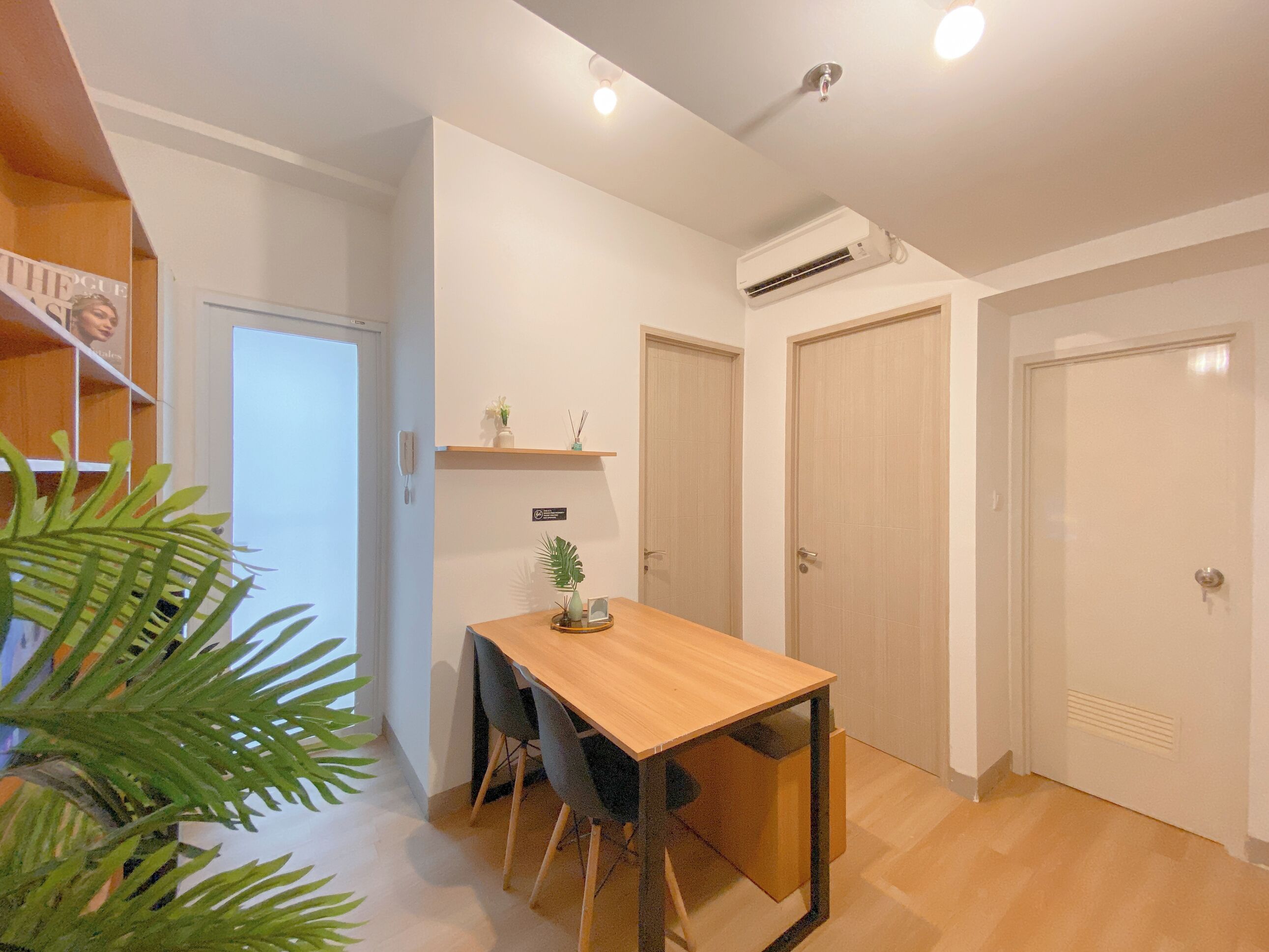 Um quarto pequeno e bem iluminado, com uma mesa de madeira, cadeiras e um vaso com planta. Há duas portas e um aparelho de ar condicionado na parede.