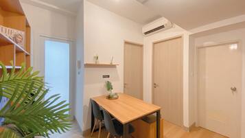 Um quarto pequeno e bem iluminado, com uma mesa de madeira, cadeiras e um vaso com planta. Há duas portas e um aparelho de ar condicionado na parede.
