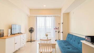 A modern living room with a blue sofa, a white dining table with a chair, a TV on a wooden stand, a standing fan, and a window with sheer curtains.