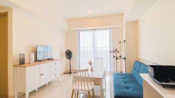 A modern living room with a sofa, a TV, a dining table with chairs, a fan, and a window with curtains.