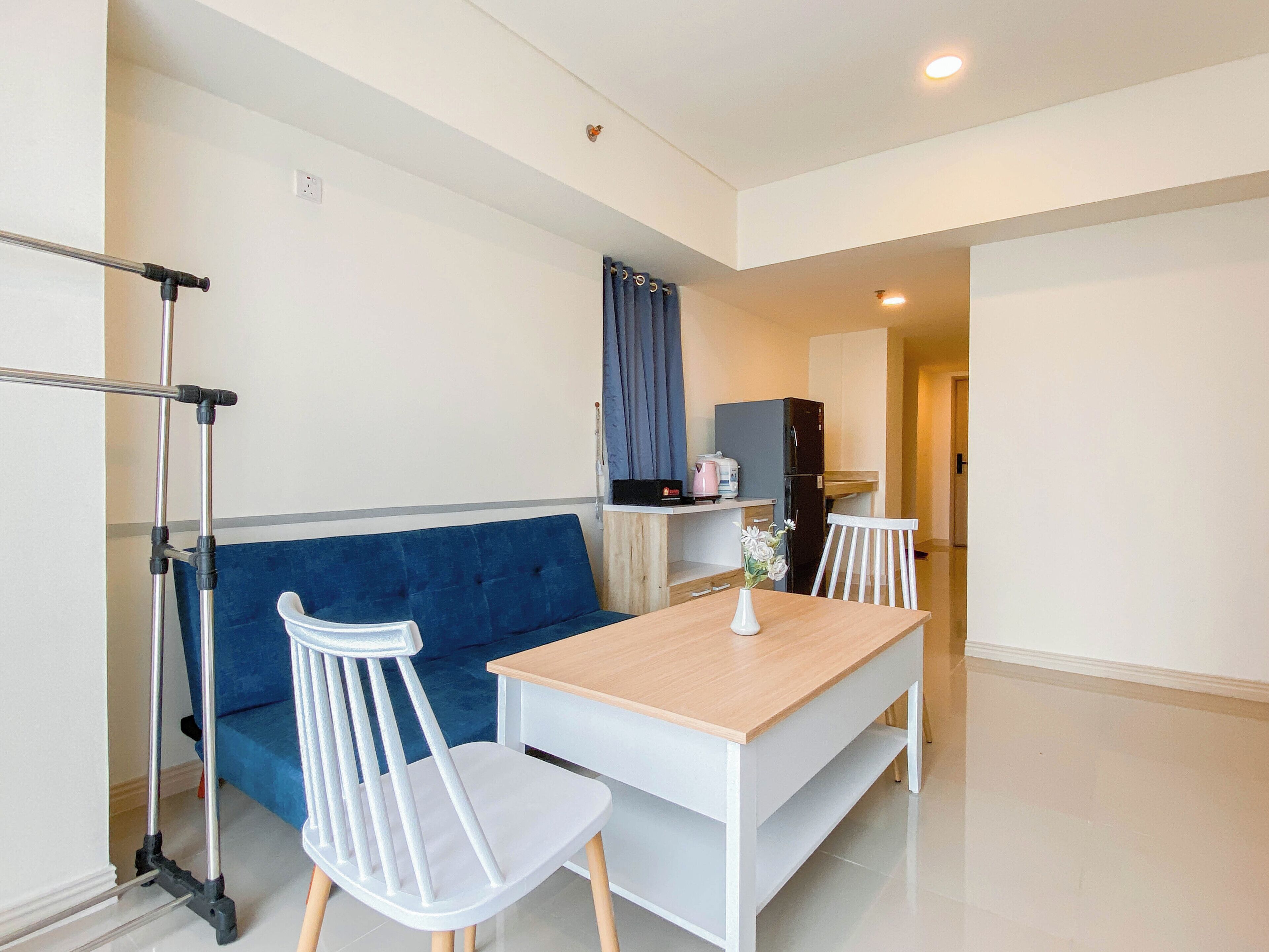 A modern, compact living space with a sofa, two white chairs, a wooden table, a kitchenette, and a ceiling-mounted exercise bar.