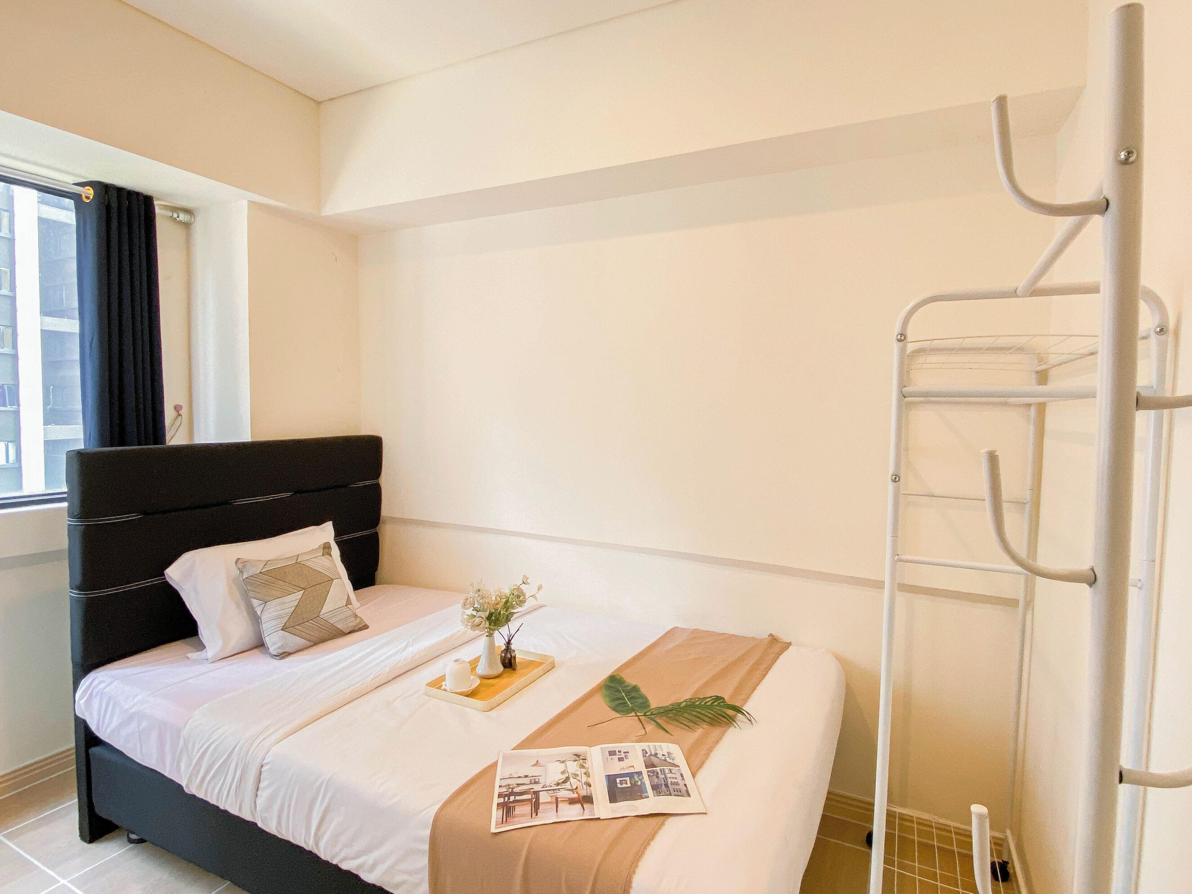 A neatly made bed with a black headboard, a window with dark curtains, and a metal drying rack against the wall.