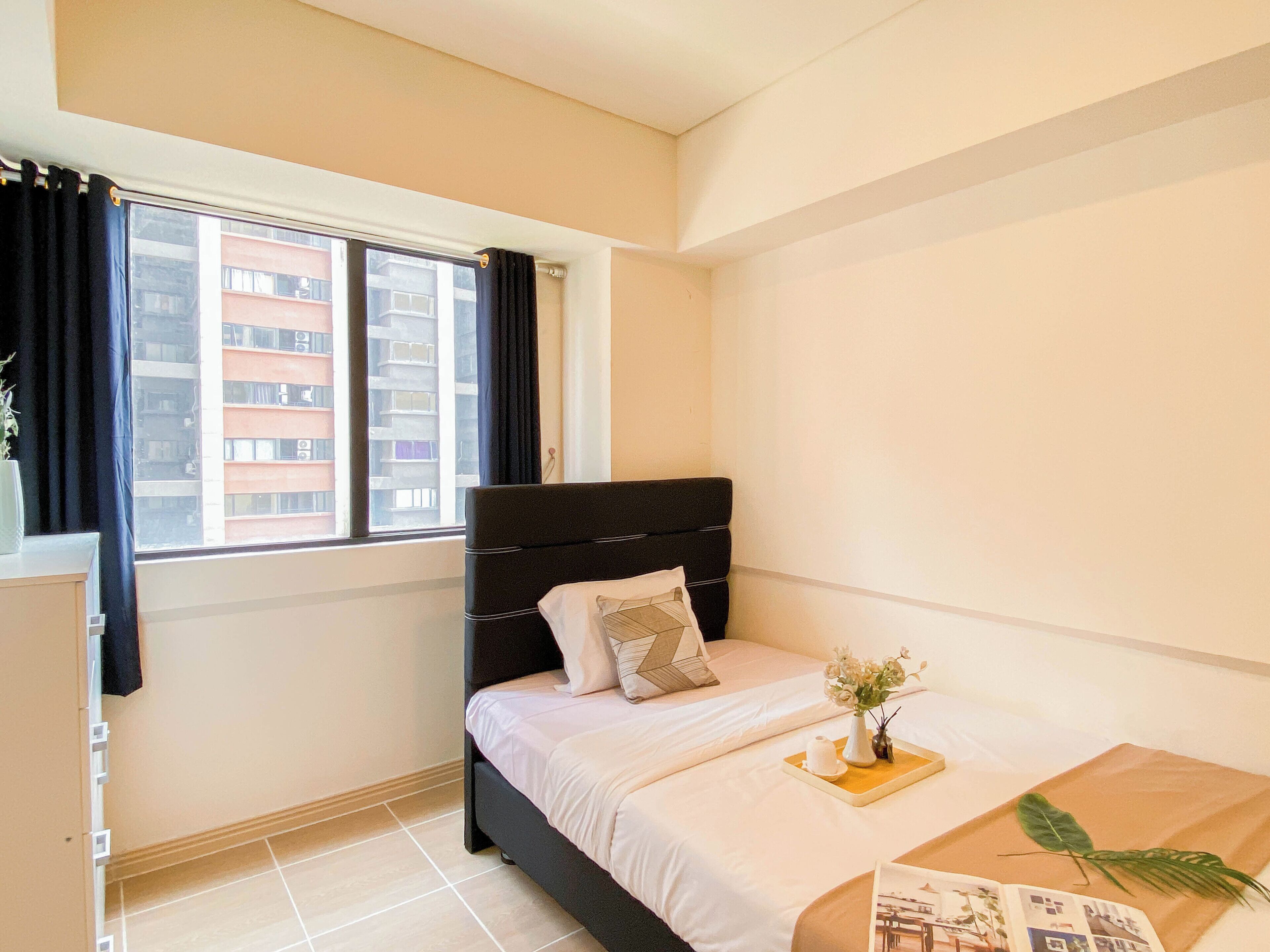 Una habitación de hotel con cama, ventana con cortinas, mesita de noche con un florero y una revista sobre la cama.