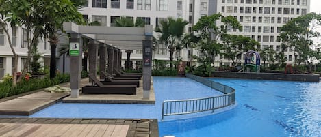 A swimming pool with a wooden deck area, surrounded by greenery and modern buildings.