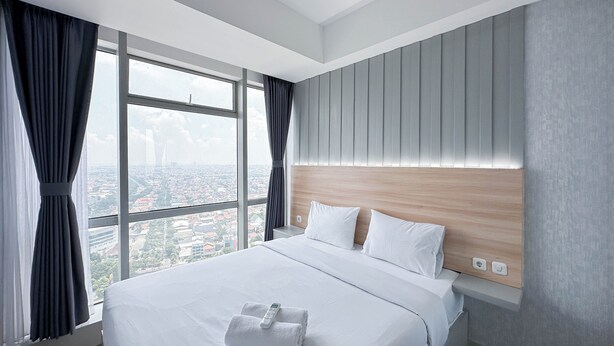 A modern hotel room with a large bed, a wooden headboard, and a city view through large windows. - Elegant And Compact 1Br At Grand Sungkono Lagoon Apartment (Surabaya)