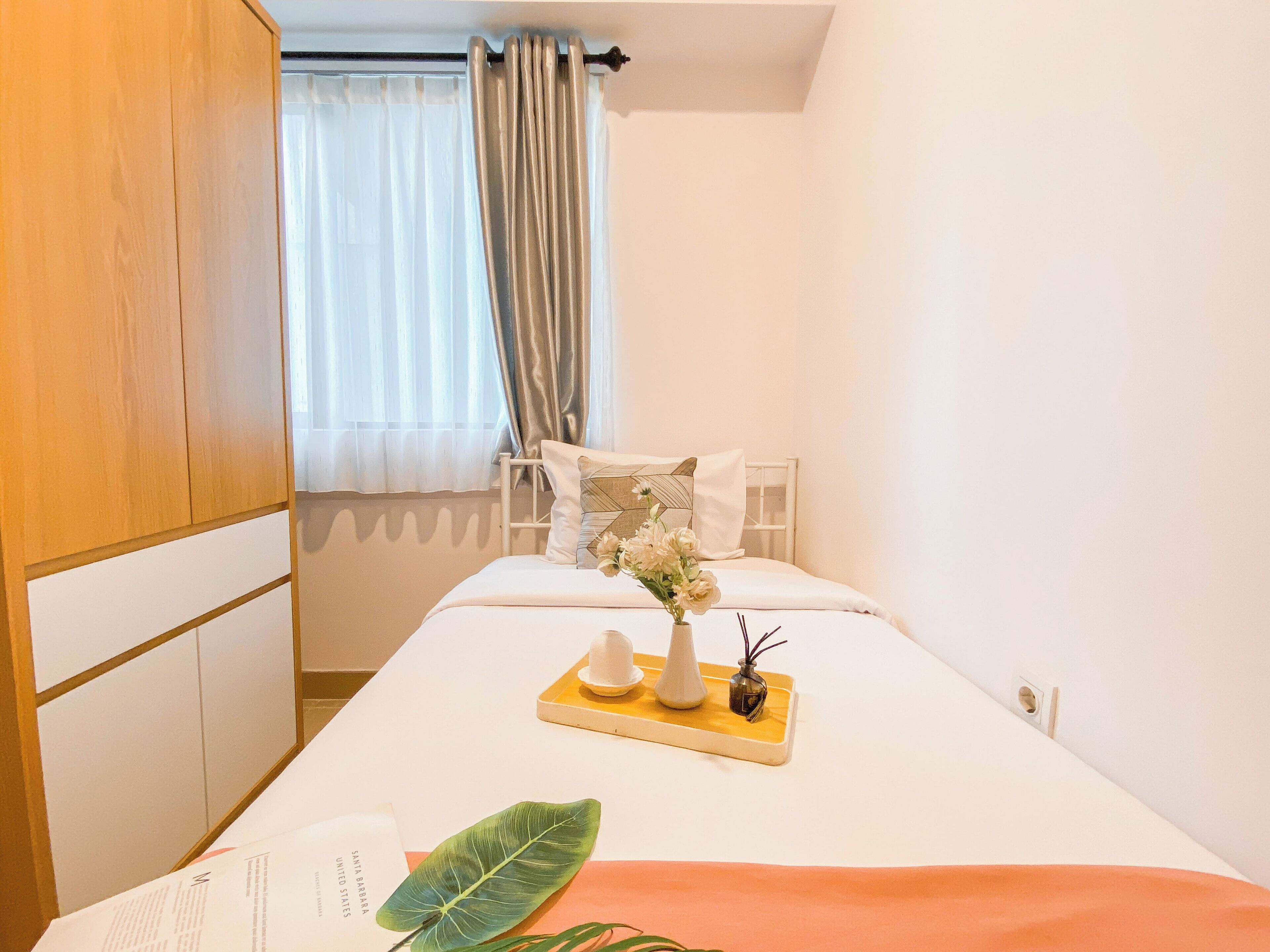Ein ordentlich eingerichtetes Hotelzimmer mit einem Bett, einem Nachttisch mit Blumen in einer Vase, einem Tablett mit Tasse und Buch sowie einem großen, grünen Blatt auf dem Bett.