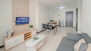 Een moderne woonkamer met een flatscreen televisie aan de muur, een grijze bank, een klein bijzettafeltje, en een eethoek met een tafel en stoelen.
