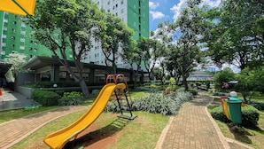 A playground with a yellow slide, surrounded by greenery and a brick pathway. - Best Price And Relaxing 1Br At Green Pramuka City Apartment (Jakarta)