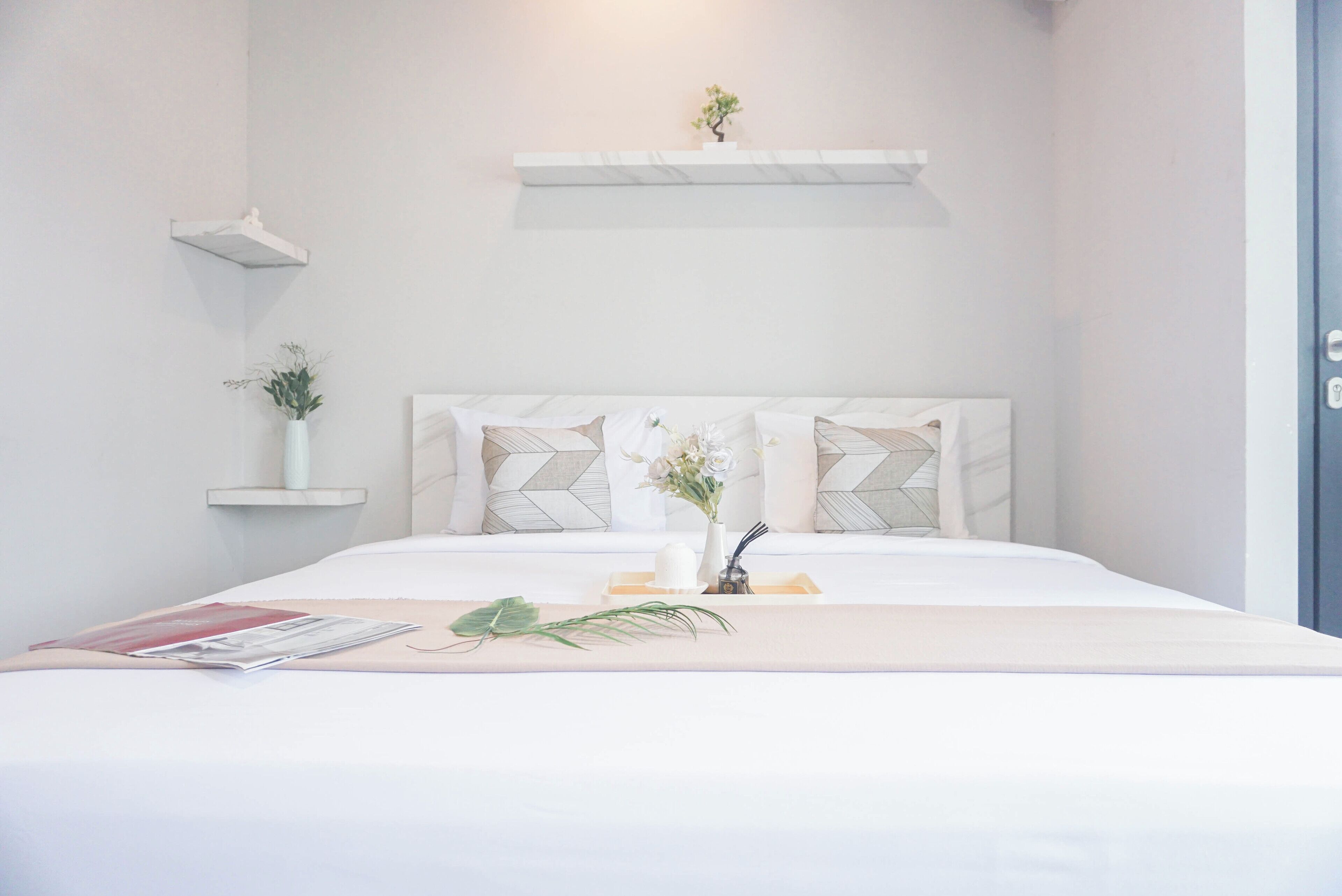 Een netjes opgemaakt bed met een wit hoofdbord, een nachtkastje met een tijdschrift en een plant, en een klein plankje met een plant en een decoratief object.