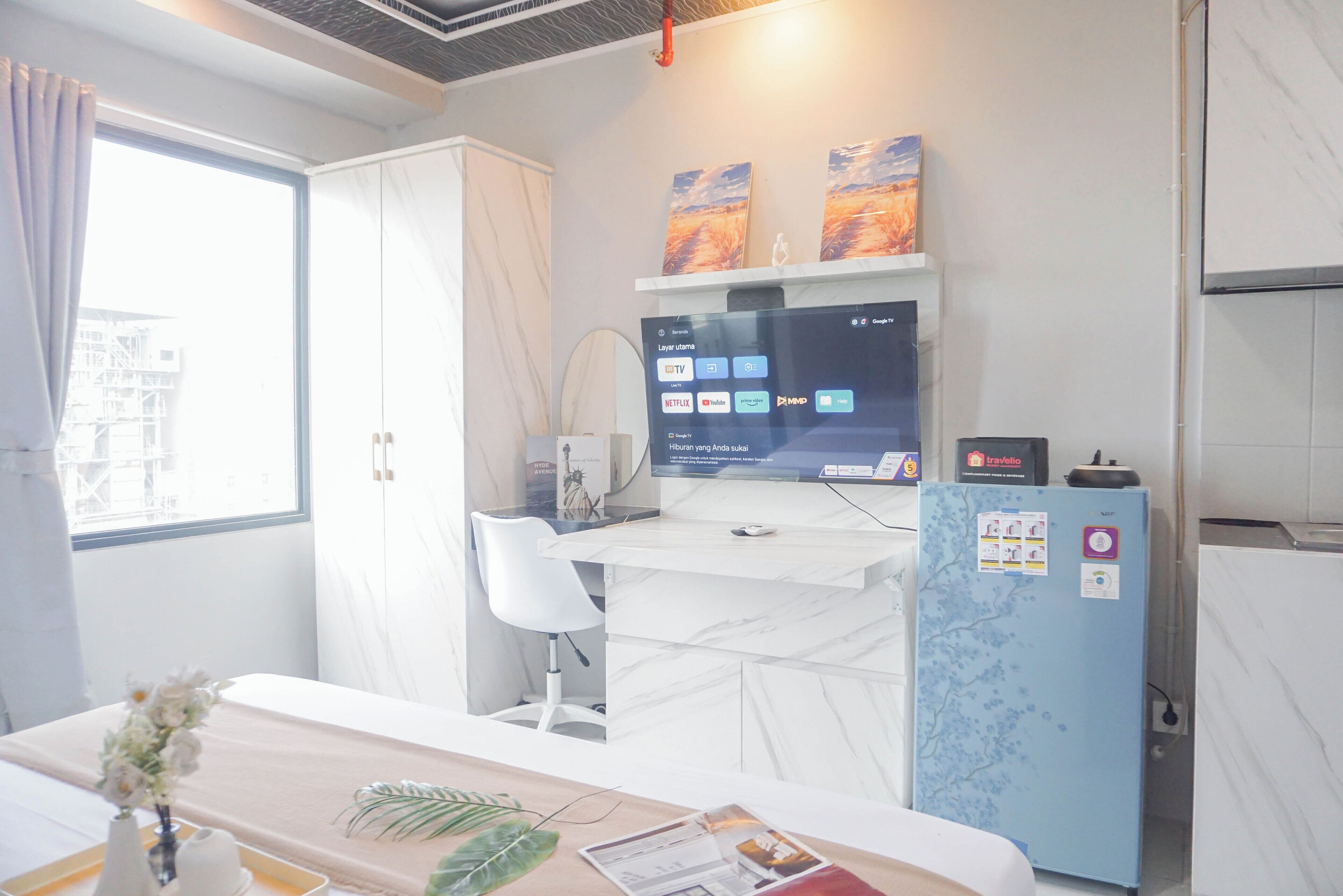 A hotel room with a bed, a desk with a computer, a blue refrigerator, and a window with curtains.