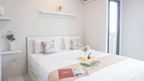 A modern bedroom with a bed, a small shelf with a plant, a bedside table with a vase and a book, and a window with a view. - Cozy And Homey Studio At Riverdale Apartment (Cikarang)