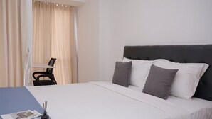 Una cama bien hecha con ropa de cama blanca y gris, un cabecero negro y una mesita azul con una revista y un portalápices.