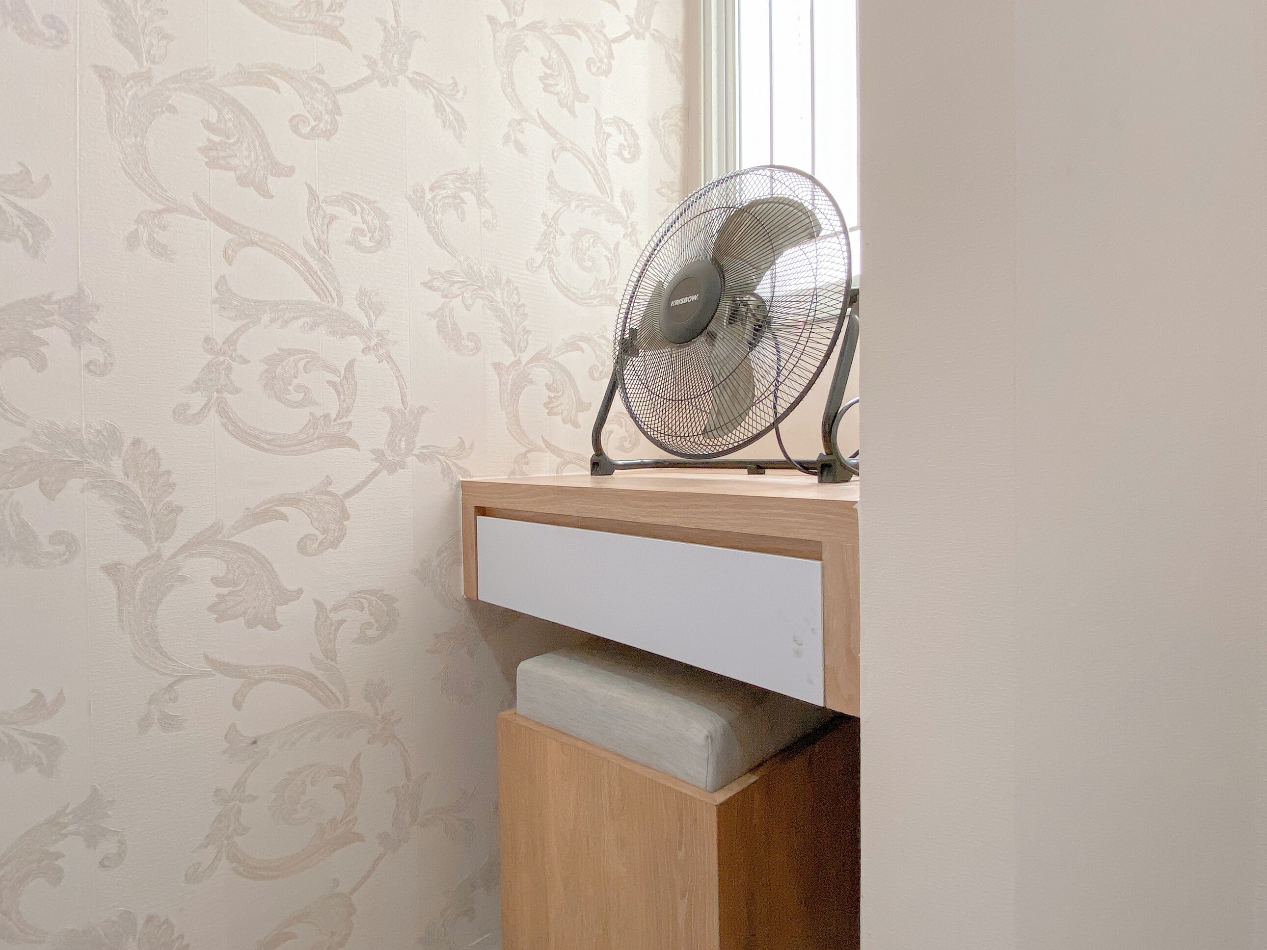 A fan on a wooden shelf in a room with patterned wallpaper.