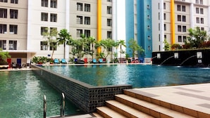 A swimming pool area with steps leading into the water, surrounded by palm trees and a multi-story building with balconies.