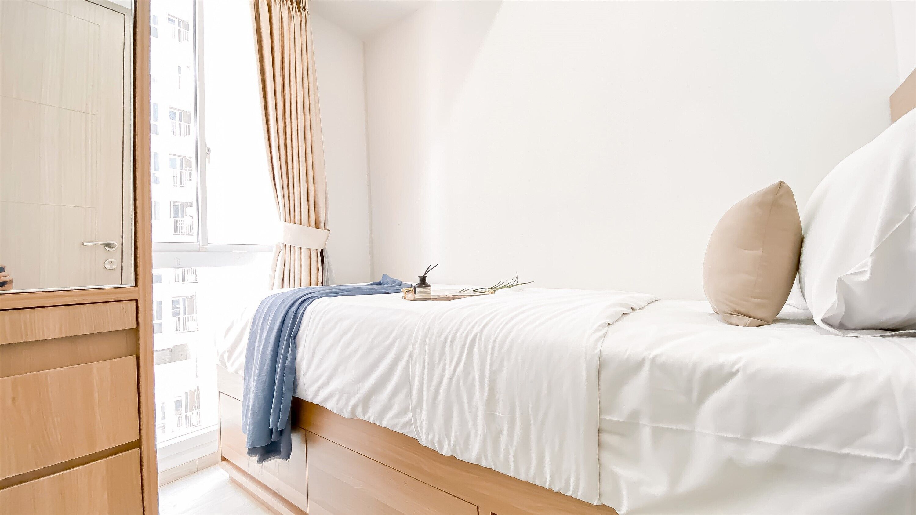 A bedroom with a wooden wardrobe, a bed with white bedding, a blue throw blanket, and a window with curtains.