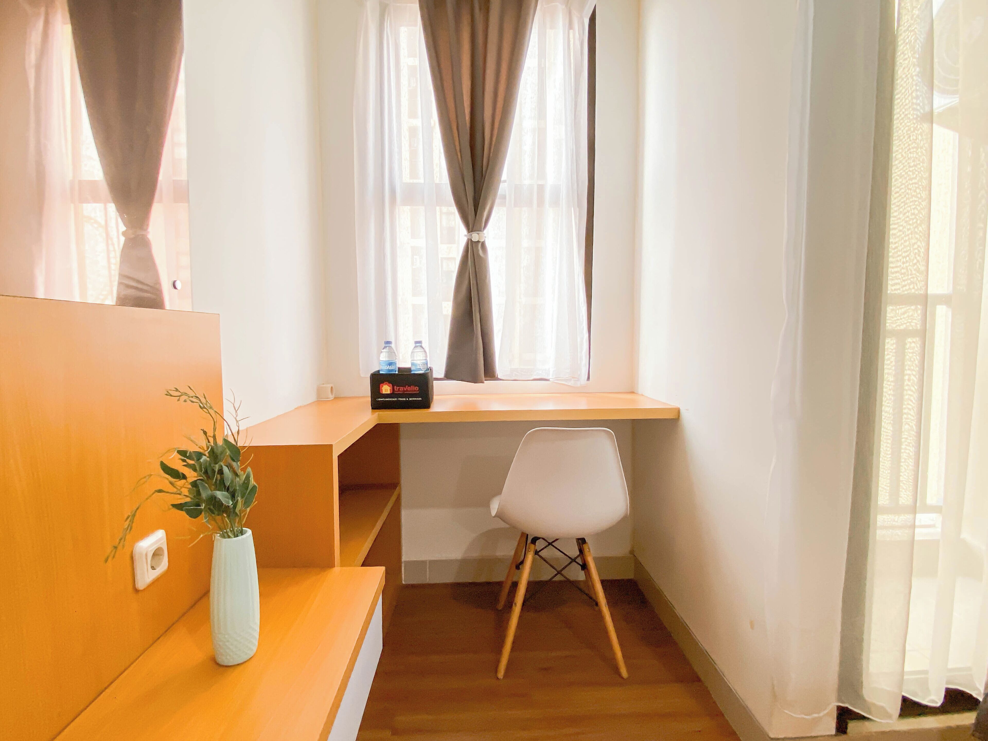 A small, well-lit room with a wooden desk, a white chair, a potted plant, and a window with curtains.