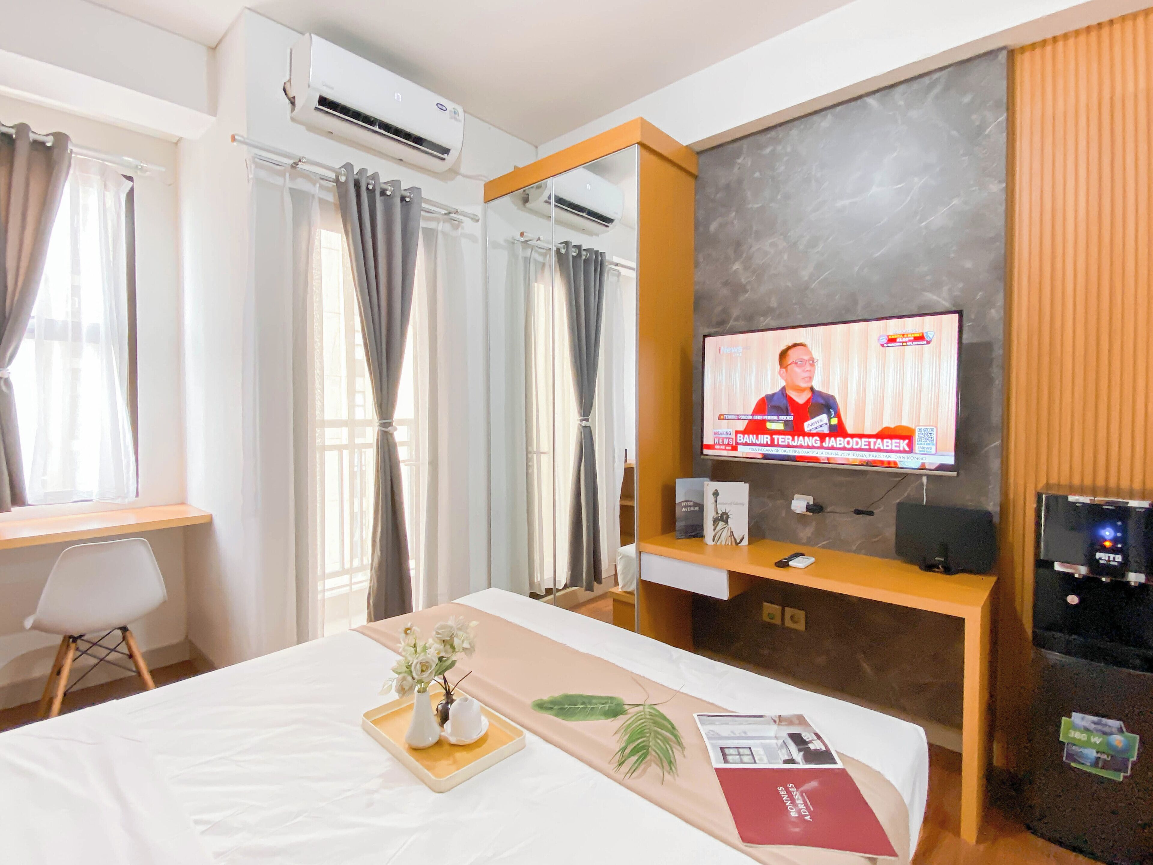 A hotel room with a bed, a TV displaying a news program, a desk with a chair, and a window with curtains.