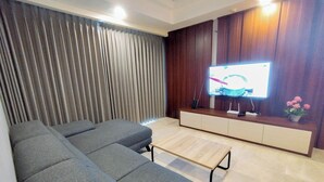 A modern living room with a grey sectional sofa, a wooden coffee table, and a flat-screen TV mounted on a wooden wall. - 2Br Luxurious And Spacious Apartment At One East Penthouse Residence (Surabaya)