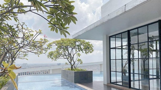 A rooftop pool with clear blue water, surrounded by potted plants and trees, and a large glass window offering a view of the city skyline. - 2Br Luxurious And Spacious Apartment At One East Penthouse Residence (Surabaya)