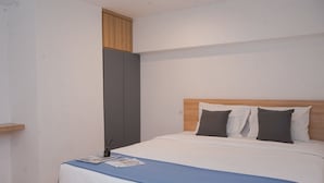 A neatly made bed with a blue bedspread and two pillows, a wooden headboard, and a small shelf with a book and a remote control.