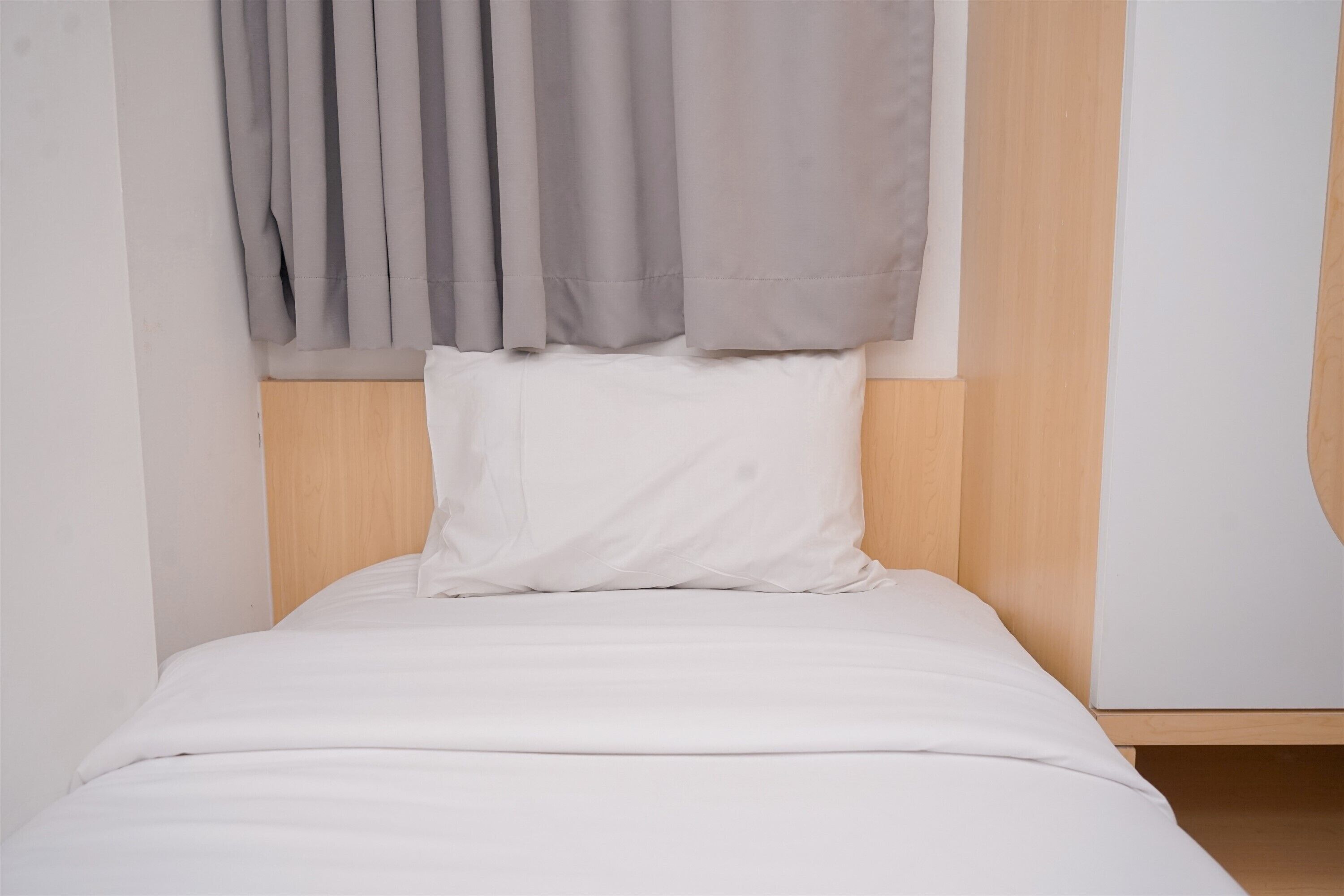 A single bed with a wooden headboard, white bedding, and a grey curtain.
