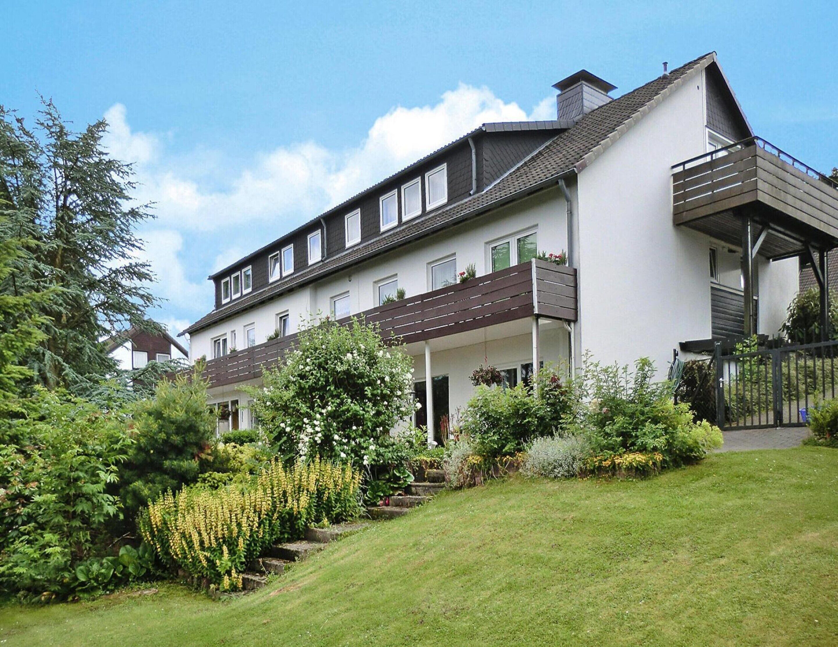 A modern two-story house with a balcony, surrounded by well-maintained gardens and a clear sky.