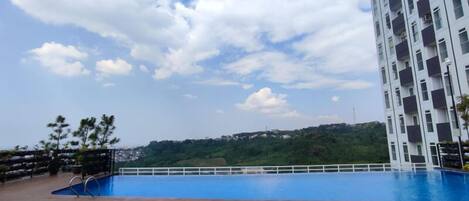 A rooftop swimming pool with a wooden deck, surrounded by a white fence, and a high-rise building in the background.