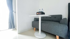 A modern room with a grey sofa, a white table with a bottle of water, and a sign indicating complimentary food and beverage. - Brand New Studio At Bassura City Apartment Near Mall (Jakarta)