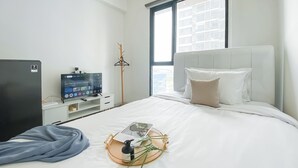 A hotel room with a bed, a TV, a window with a view of buildings, and a bedside table with a bottle and glasses.