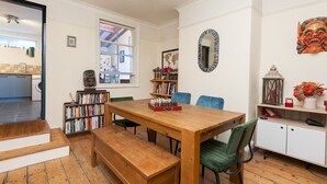 Un coin repas avec une table en bois, des chaises bleues et un banc. On y trouve une bibliothèque remplie de livres et un miroir décoratif accroché au mur.
