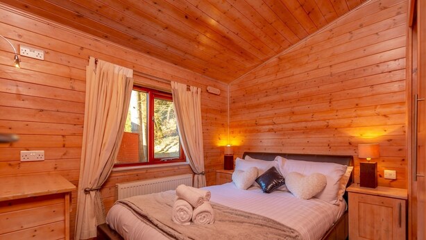 A cozy wooden cabin bedroom with a bed, bedside lamps, and a window with curtains. - Astbury Falls Escape – A Tranquil Stay in Nature - Pass the Keys (Bridgnorth)