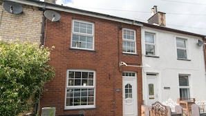 A two-story brick house with a white door and windows. There is a satellite dish on the roof, and a small garden area with a potted plant.