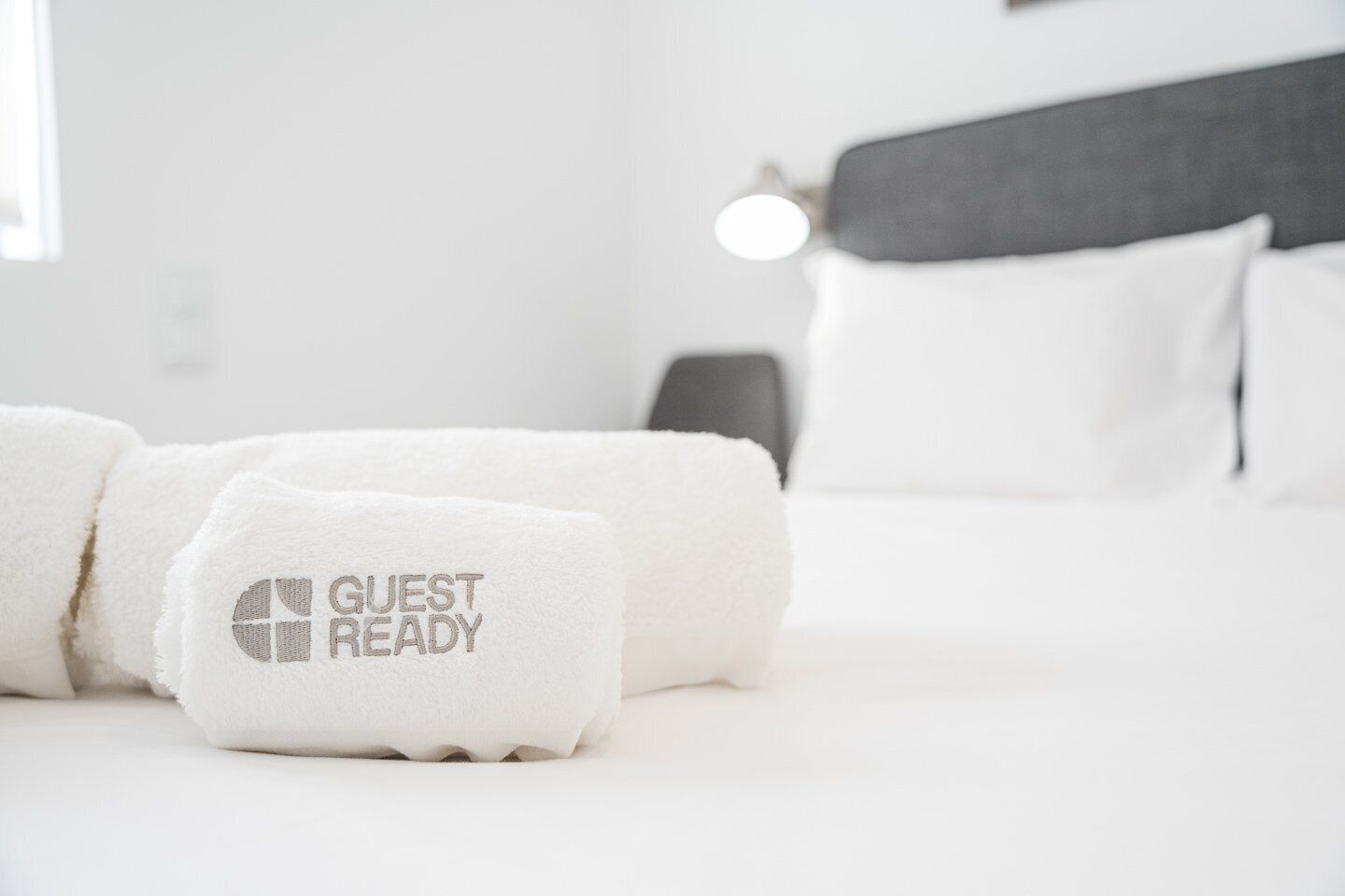 A neatly made bed with a white comforter and two rolled towels with the text "GUEST READY" on them.