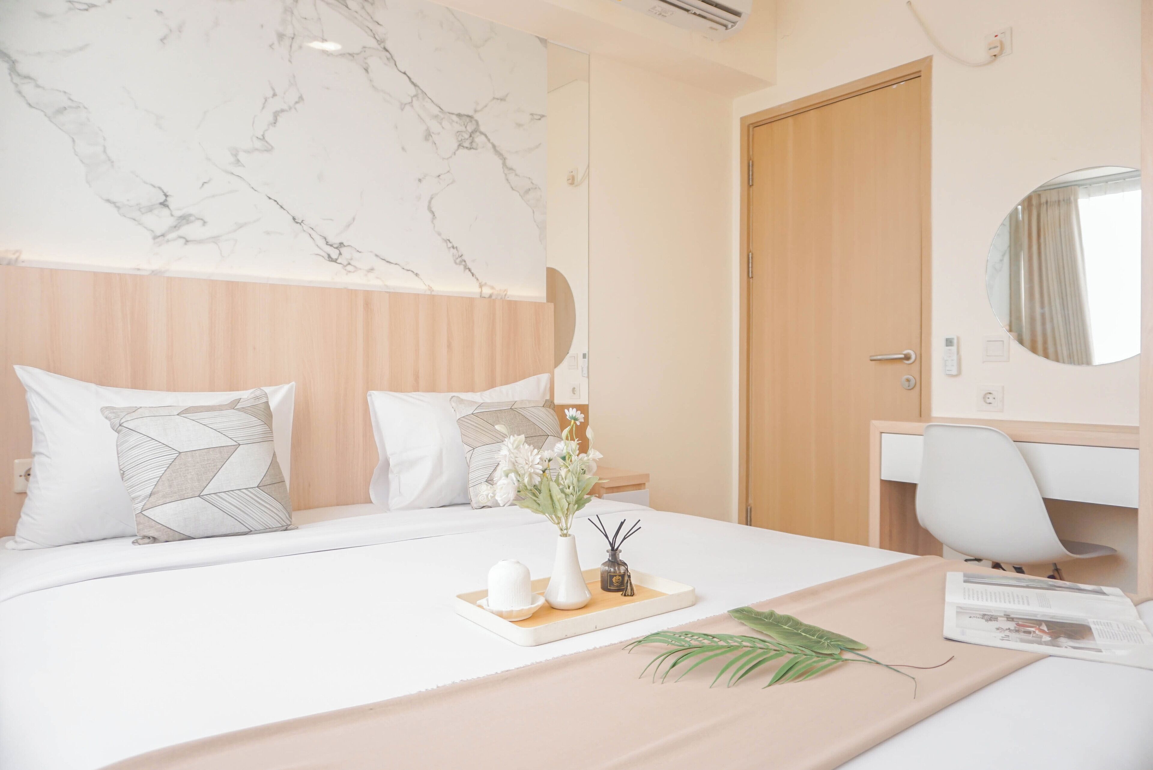 A modern hotel room with a marble headboard, a wooden door, a white desk, and a bed with a tray of toiletries.