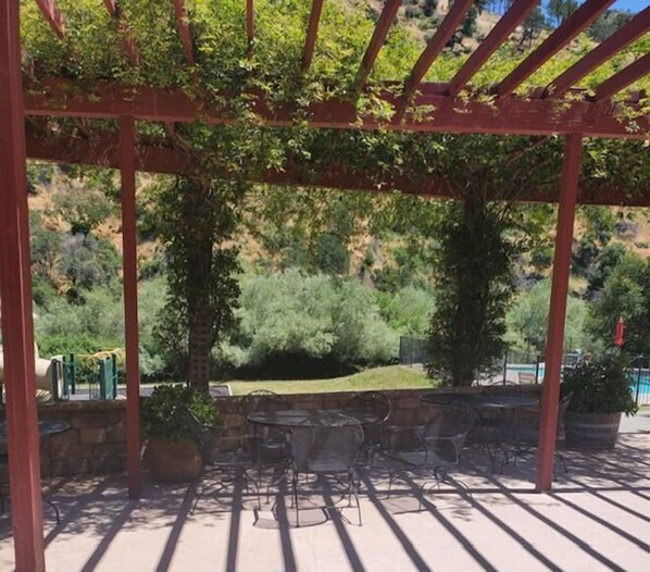 Un patio exterior con pérgola, mesas y sillas, con vistas a un jardín ajardinado y colinas.