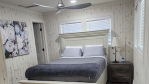 Un dormitorio con cama, mesita de noche, ventilador de techo y ventana con persianas.