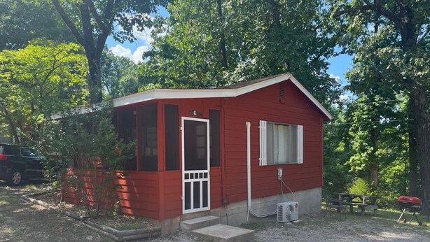 A small red cabin with a white door and windows, a gravel driveway, and a picnic table nearby.