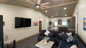 A modern living room with a flat-screen TV, a ceiling fan, and a dining area with a kitchen in the background. - Boater's Bliss at Fish & Fiddle Family Resort & Extended Stay (Mountain Home)
