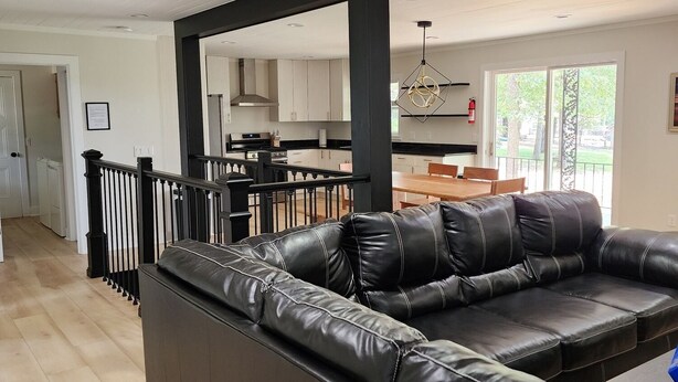 A modern living room with a black leather sectional sofa, a staircase with black railings, and a kitchen in the background. - Secluded Hideaway at Fish & Fiddle Family Resort & Extended Stay (Mountain Home)
