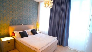 Una cama bien hecha con almohadas, una lámpara de noche y una mesita de noche en una habitación con papel tapiz estampado y un ventanal con cortinas.
