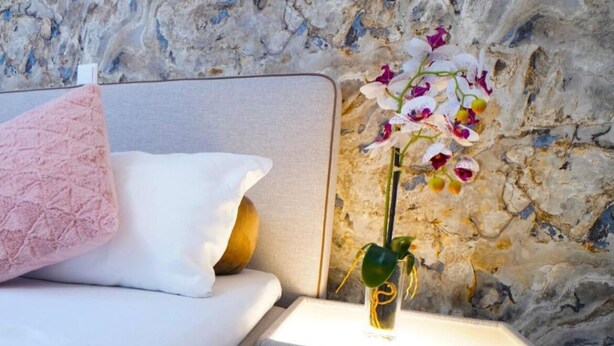 Un dormitorio con cama, almohadas, una mesita de noche con un florero y una pared de piedra.