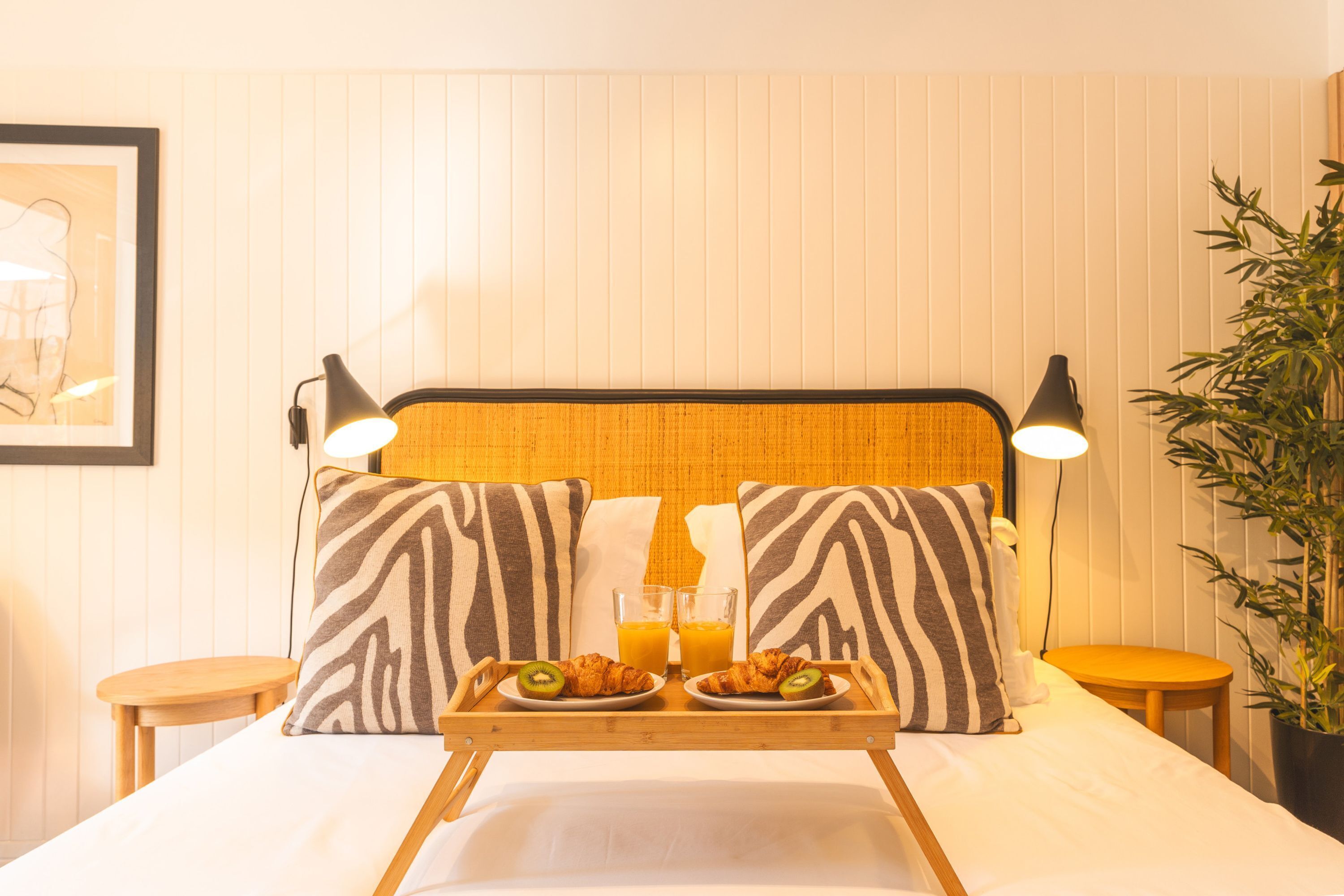 Habitación de hotel con una cama, una bandeja con croissants y zumo de naranja, dos lámparas de noche y una planta en maceta.